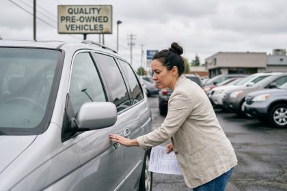 Femme examinant une voiture familiale d'occasion dans un parking de concessionnaire, tenant une liste de vérification